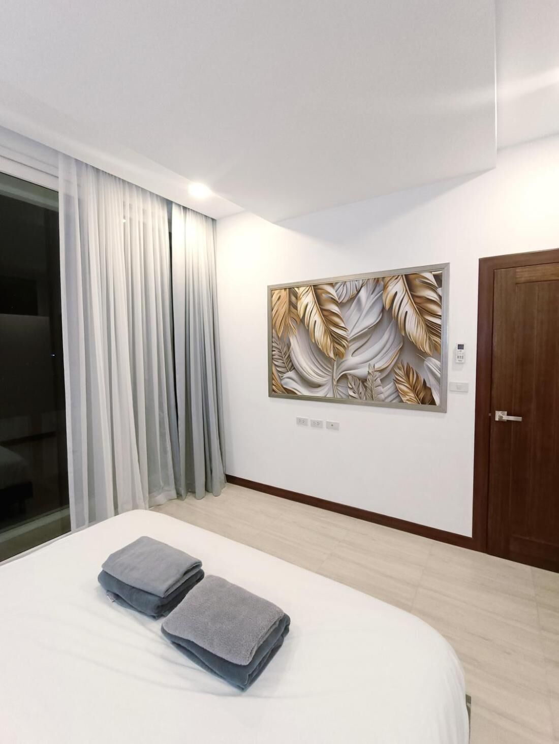 A hotel room with a bed, a large window with curtains, a wall-mounted artwork, and a wooden door.