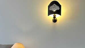 Una mesita de noche de madera con una lámpara y un florero, una silla estampada y un aplique de pared.
