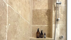 Zona de ducha con azulejos color beige, cabezal de ducha de mano y dos botellas de jabón en un estante.