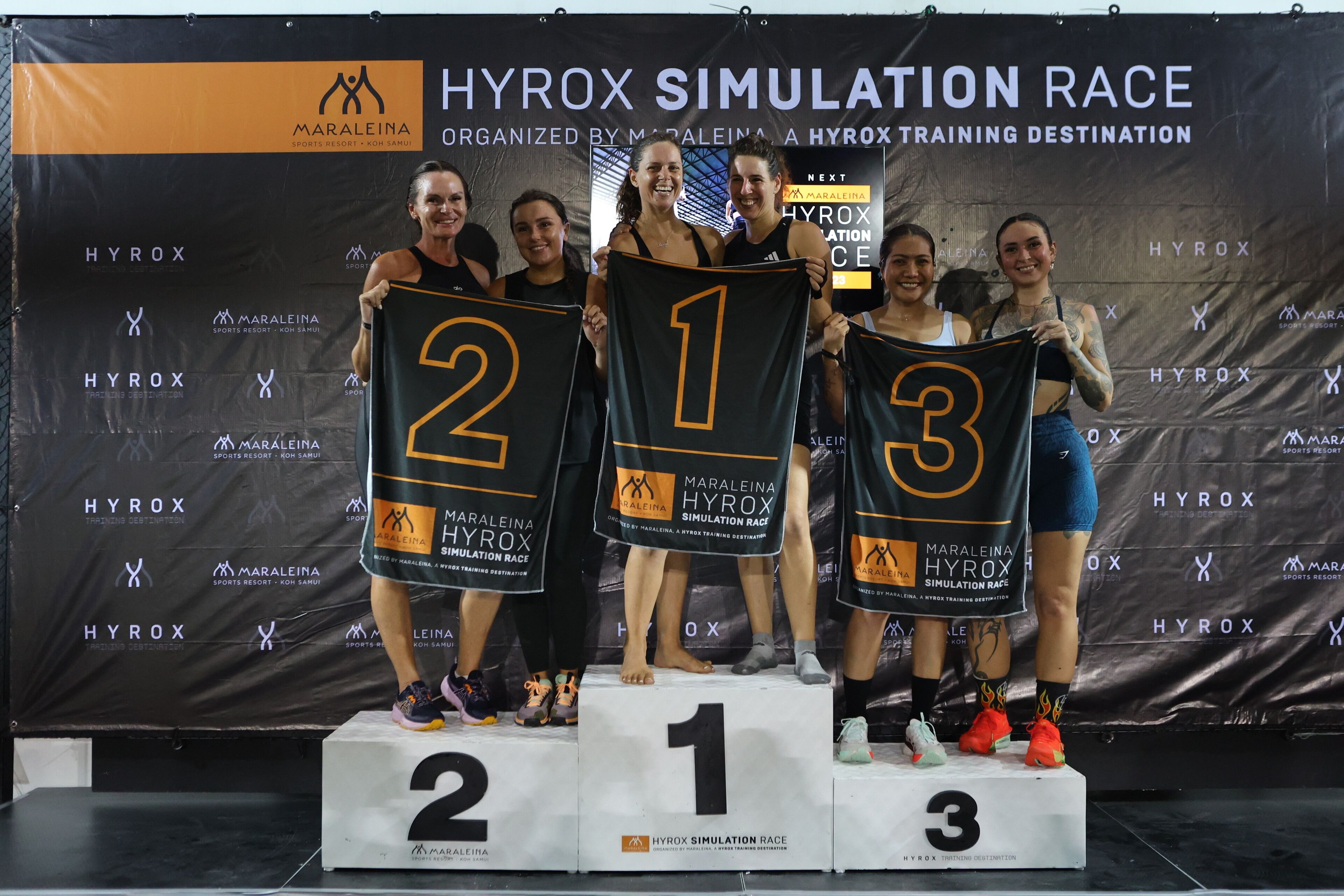 Pódio com três pessoas usando camisas numeradas, em um palco com um painel de fundo mostrando o nome do evento e patrocinadores.
