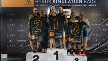 Pódio com três pessoas usando camisas numeradas, em um palco com um painel de fundo mostrando o nome do evento e patrocinadores.