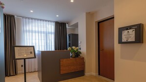 A hotel reception area with a wooden desk, a framed certificate on the wall, and a large window with curtains. - Luna Stay (Trieste)