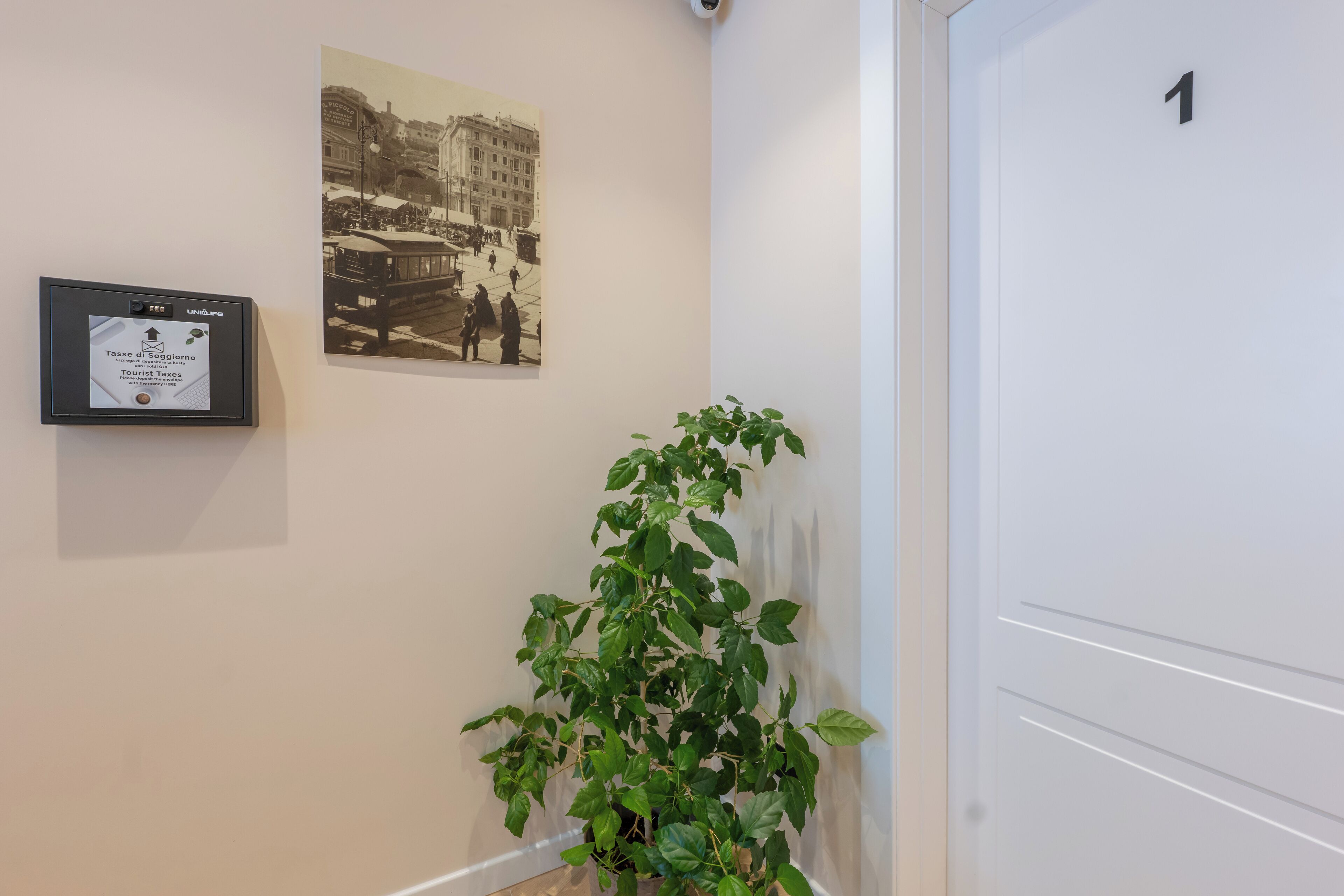 Un couloir orné d’une photographie historique encadrée, d’un panneau d’information sur la taxe de séjour et d’une plante en pot.