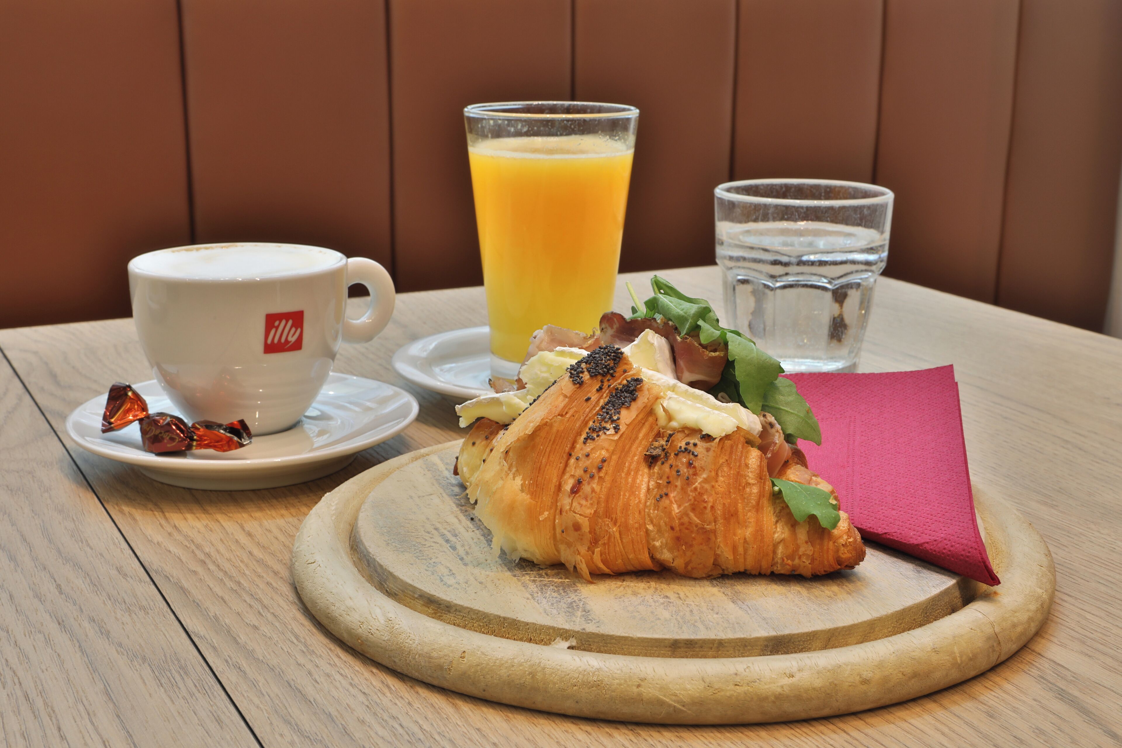 Un desayuno con un croissant, un vaso de jugo de naranja, una taza de café y un vaso de agua sobre una mesa de madera.