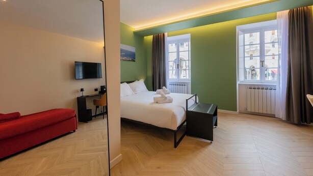 A modern hotel room with a bed, a red sofa, a desk with a chair, a TV, and large windows with curtains. - Luna Stay (Trieste)