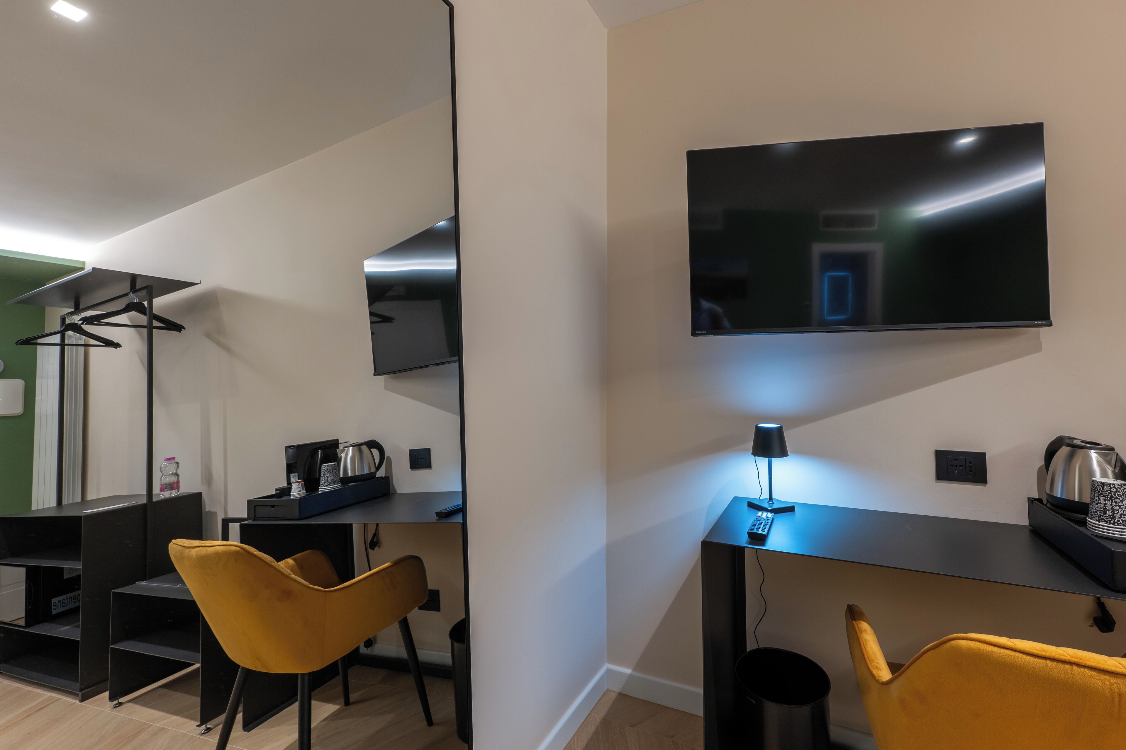 Ein modernes Hotelzimmer mit einem wandmontierten Flachbildfernseher, einem Schreibtisch mit Lampe, einer Kaffeemaschine und einem Sessel.