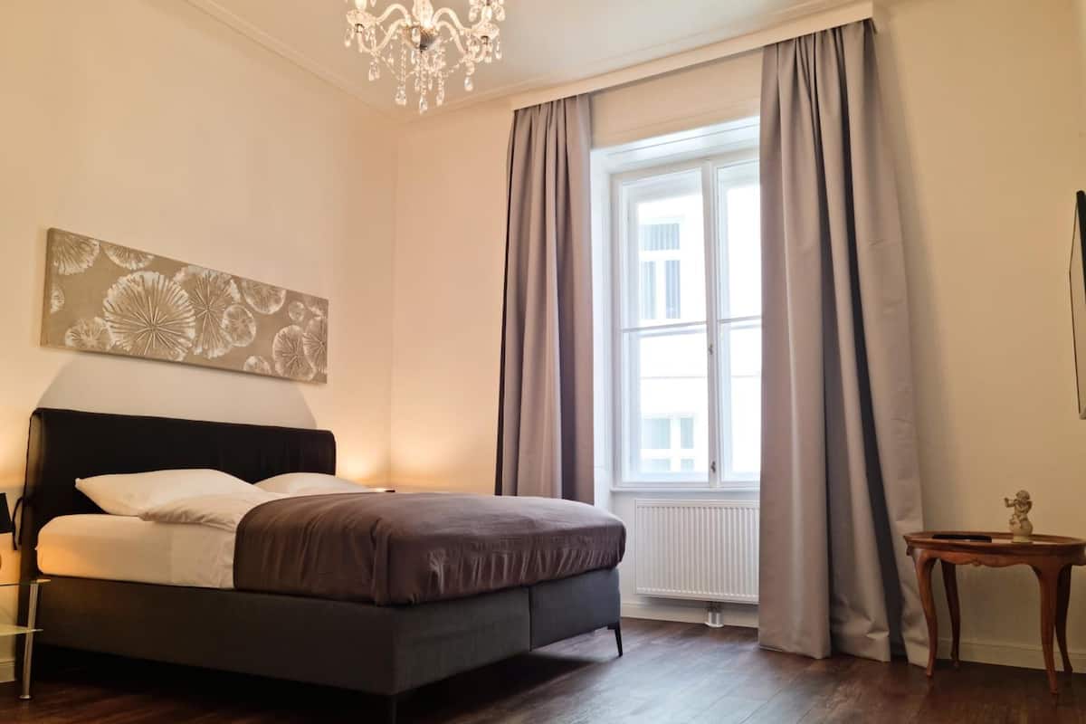 Ein Schlafzimmer mit einem Bett, einer Chandelier, einem Fenster mit Vorhängen und einem kleinen Tisch mit einer Statue.