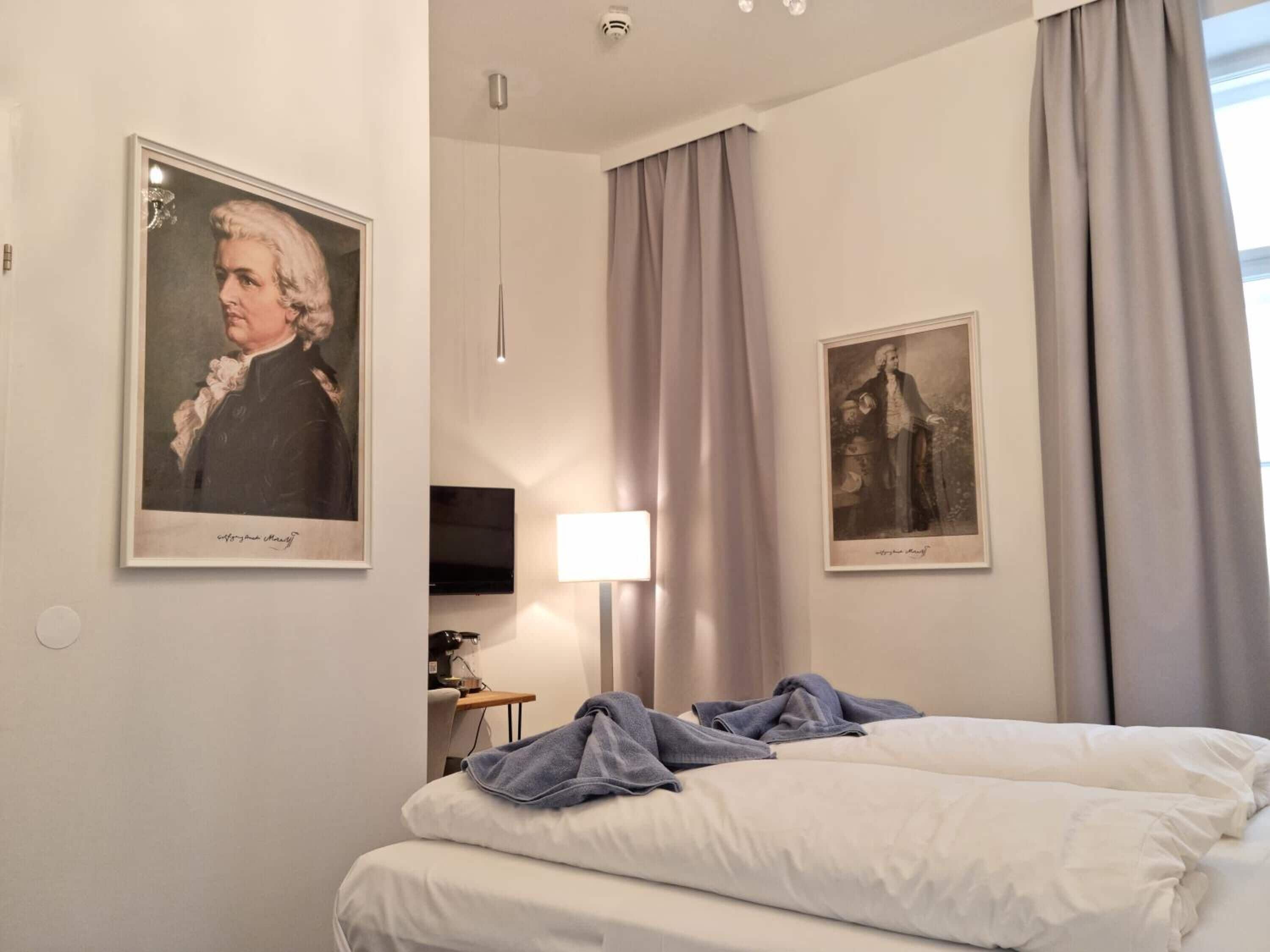 A hotel room with a bed, a desk, a television, and two framed portraits on the wall.