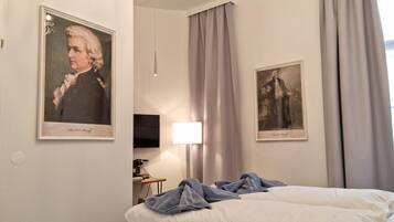 A hotel room with a bed, a desk, a television, and two framed portraits on the wall.