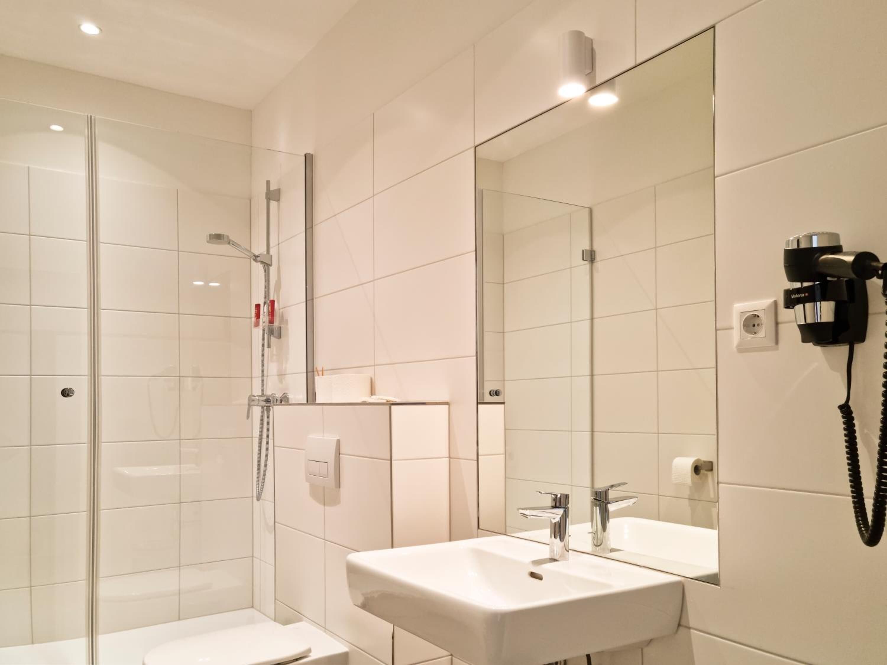 Ein modernes Badezimmer mit Duschkabine aus Glas, einem weißen Waschbecken mit Spiegel und einem an der Wand befestigten Föhn.