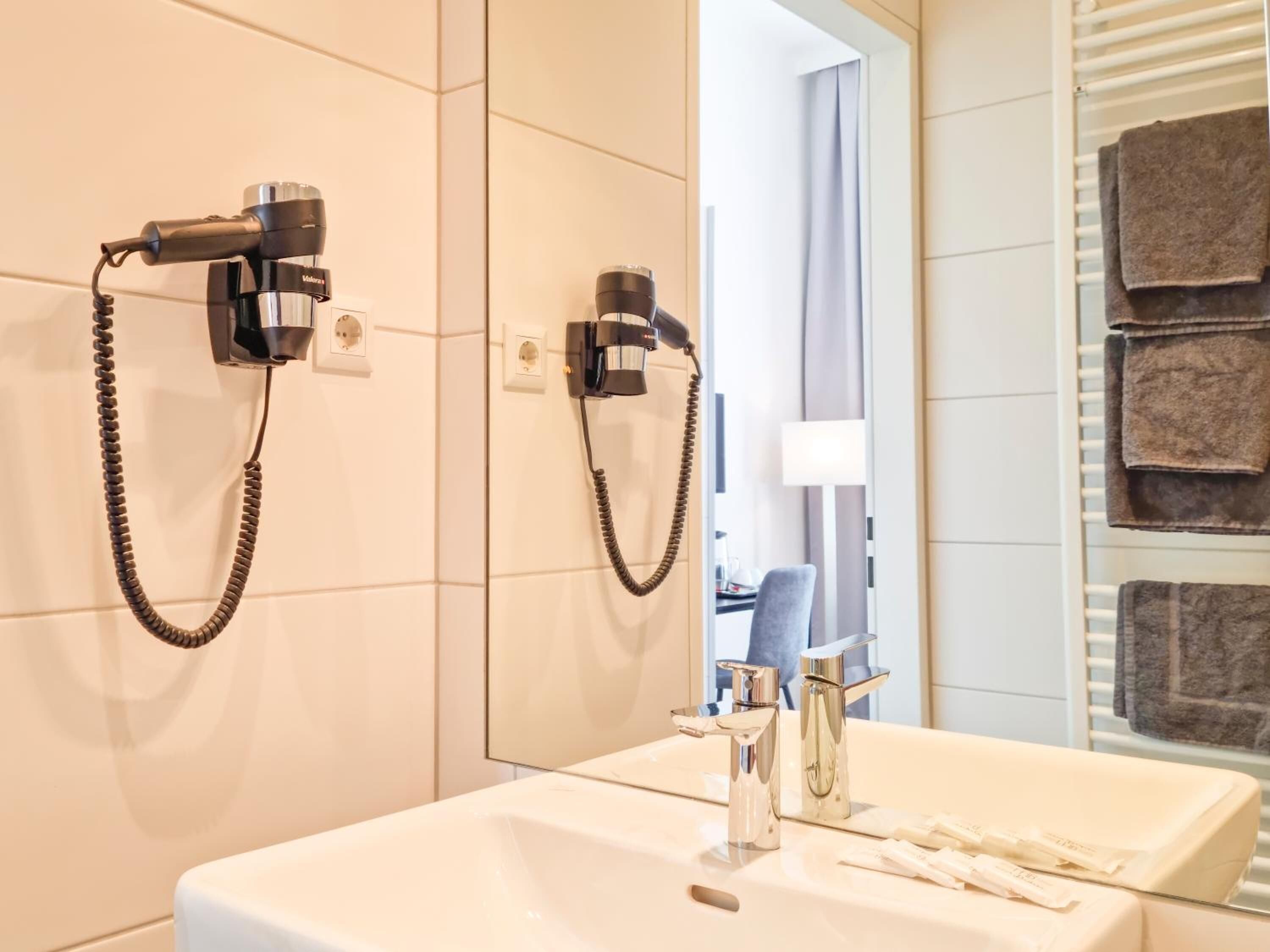 Un baño con lavamanos, dos secadores de pelo montados en la pared, un perchero con toallas dobladas y un espejo.