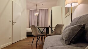 A modern hotel room with a wooden floor, a grey sofa, a dining table with chairs, a chandelier, and a lamp.