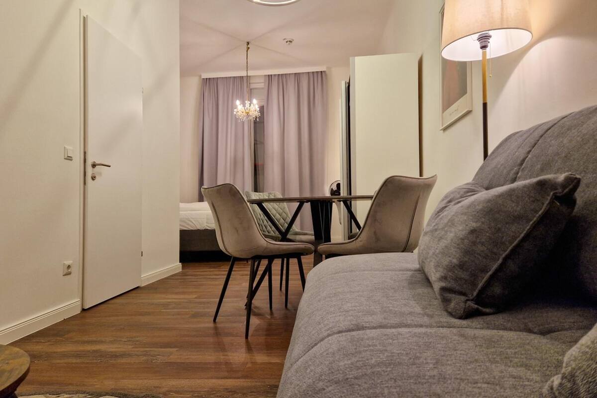 Ein modernes Hotelzimmer mit Holzboden, einem grauen Sofa, einem Esstisch mit Stühlen, einer Kristallleuchter und einer Lampe.