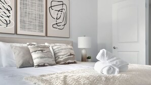 Ein ordentlich bezogenes Bett mit einem aufgerollten Handtuch, einer strukturierten Tagesdecke und gestreiften Kissen. Darüber befinden sich abstrakte Wandbilder.