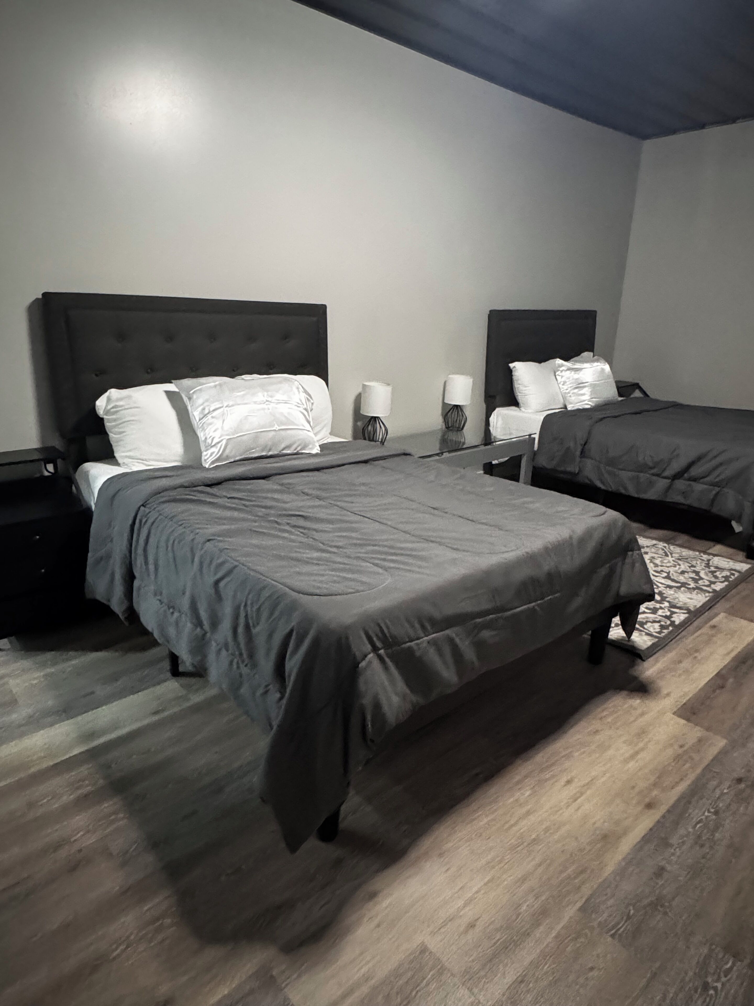 Quarto de hotel com duas camas, cada uma com cabeceira, mesa de cabeceira com abajur e tapete estampado no chão.