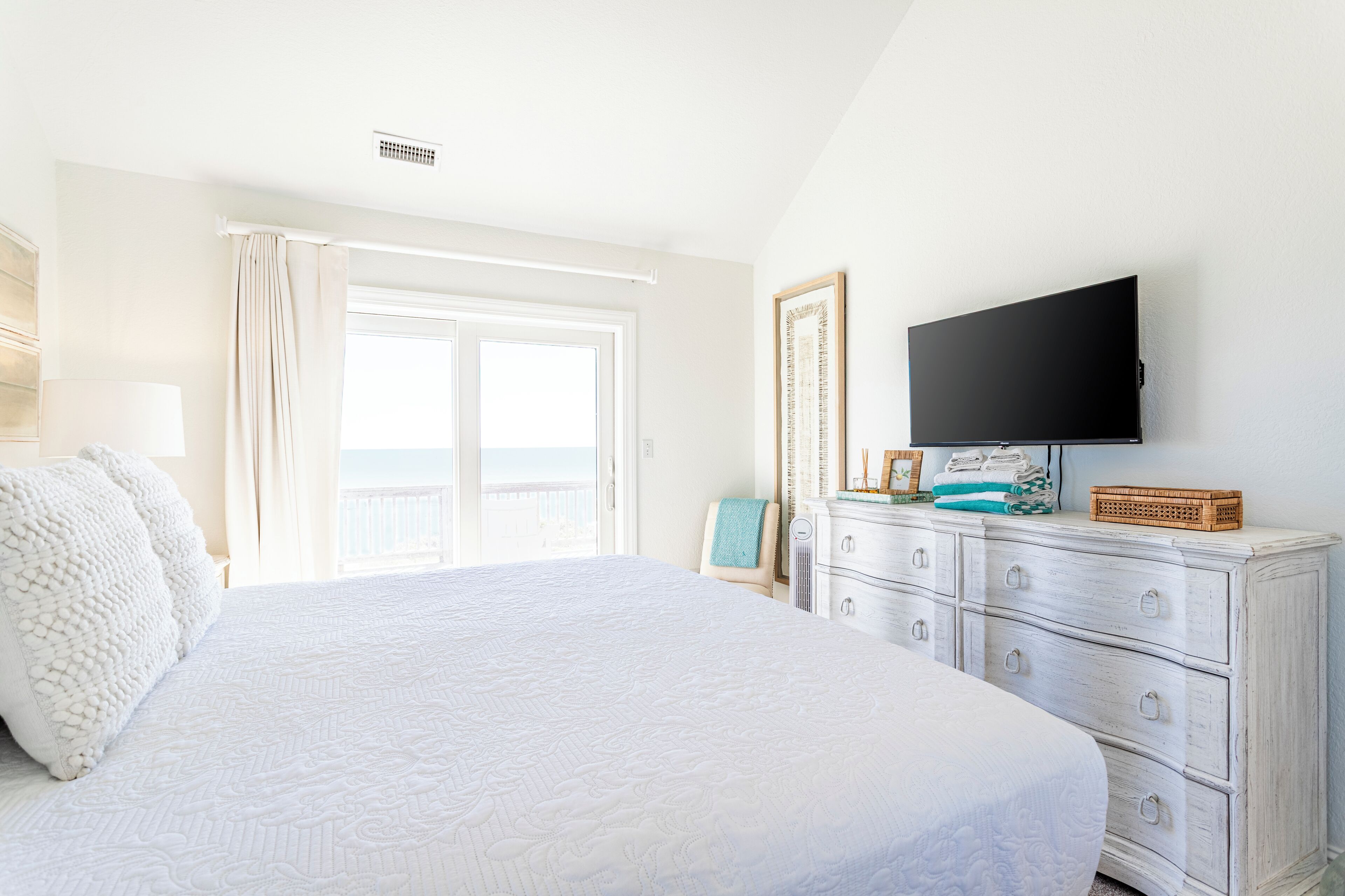 A bedroom with a large bed, a dresser, a flat-screen TV, and a window with curtains.