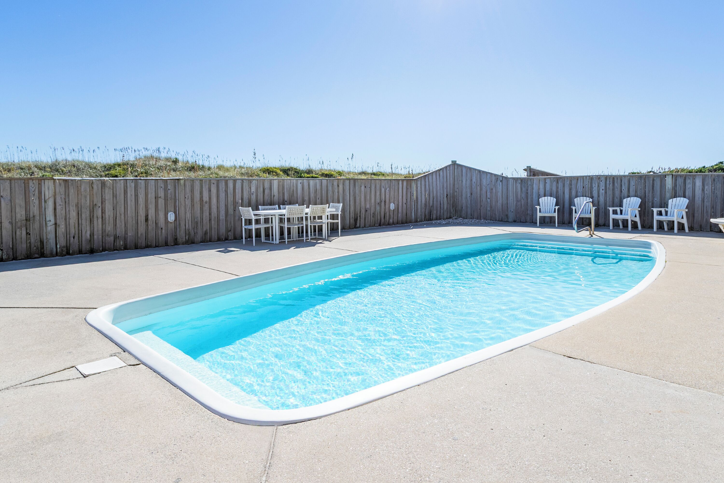 Ein rechteckiger Pool mit Holzzaun und weißen Stühlen darum herum.
