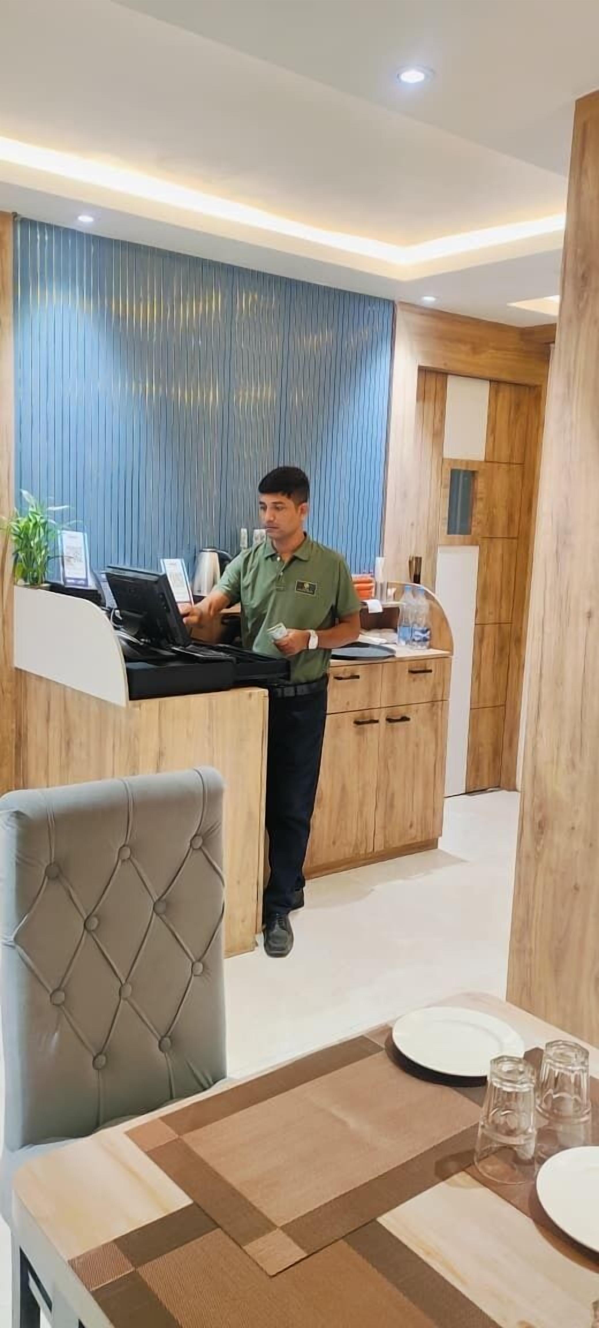 Un espace d’accueil d’hôtel au design moderne, comprenant un comptoir d’accueil en bois, un ordinateur et un coin salon avec un fauteuil capitonné et une table préparée pour deux personnes.