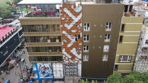 A multi-story building with a distinctive geometric facade pattern, a rooftop terrace, and a visible commercial area with shops and parked vehicles. - Hotel Green View (Berhampore)