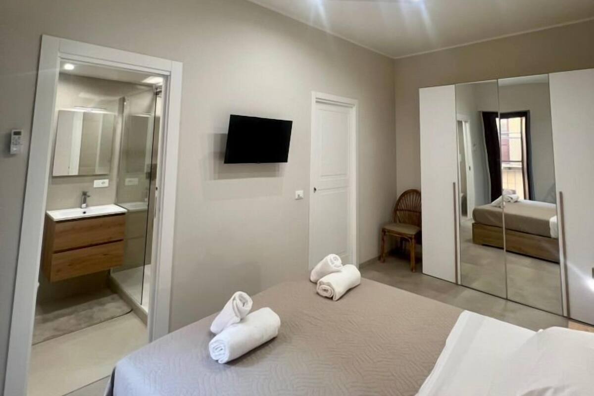 A modern bedroom with a bed, a TV mounted on the wall, a mirror, and a bathroom visible through an open door.