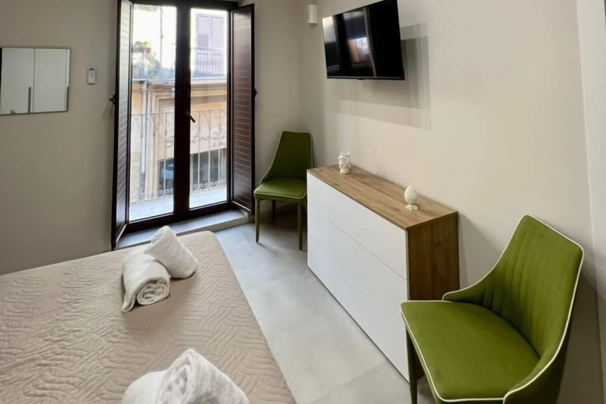 A modern hotel room with a bed, a TV, a green chair, and a sliding glass door to the balcony.