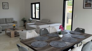 Ein modernes Wohnzimmer mit Essbereich, grauen Sofas, einem Couchtisch mit Blume und Blick durch ein Fenster.