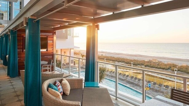 A balcony with a view of the beach, a pool, and a seating area with cushions. - Ocean Enclave July 4th weekend. Beachfront hotel. Partial Ocean View 1 bedroom. (Myrtle Beach)