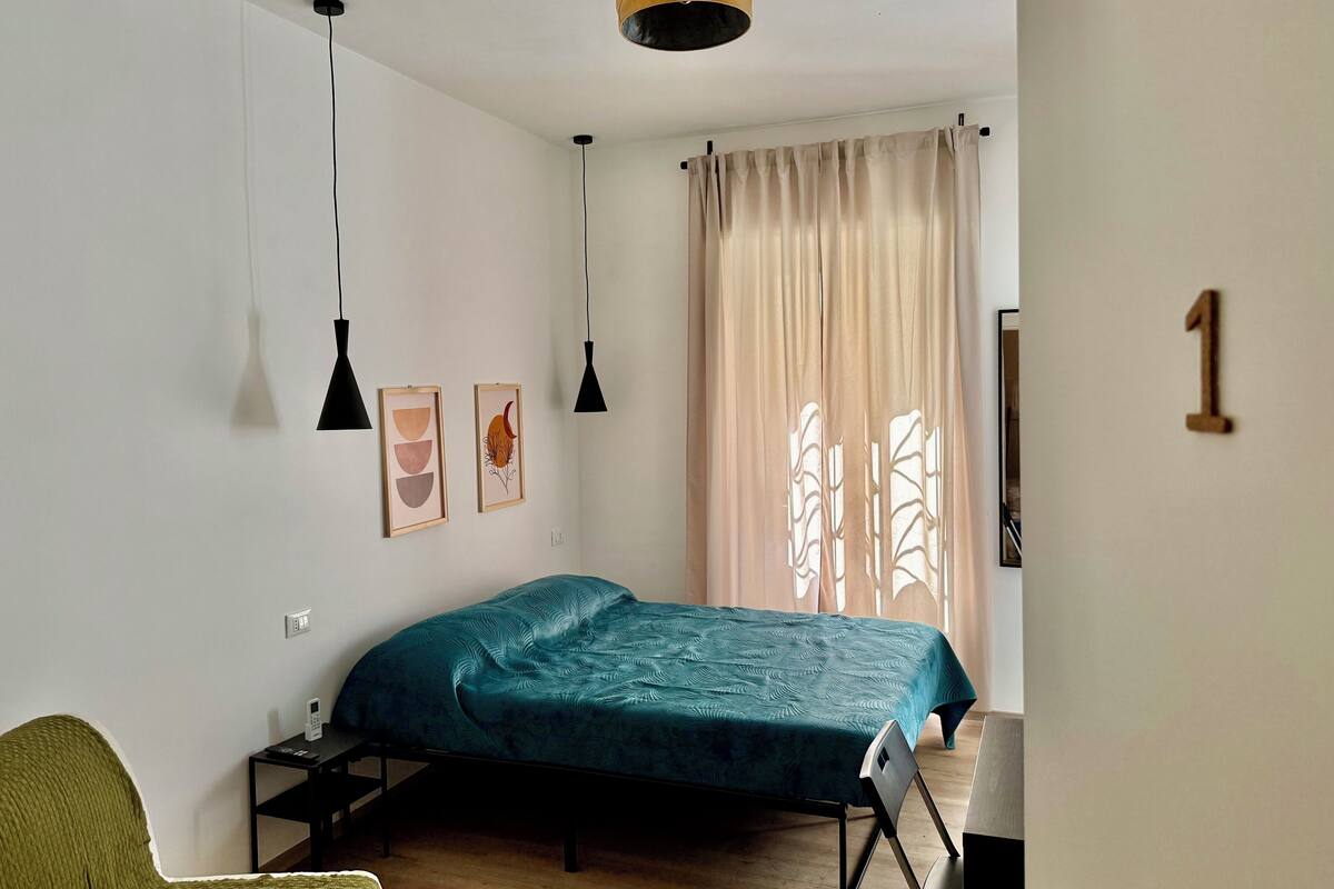 A bedroom with a bed, two pendant lights, a wall-mounted number, and a window with curtains.