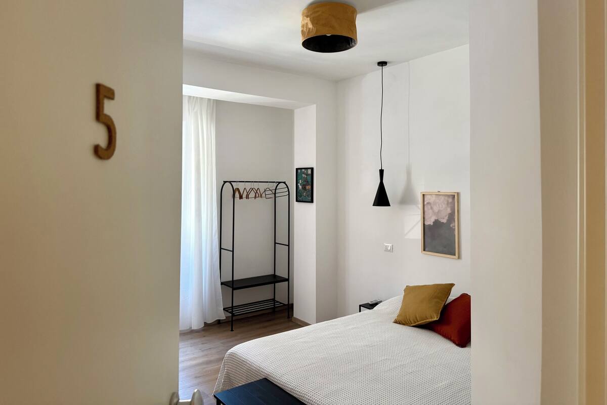 A bedroom with a bed, a hanging lamp, a wall-mounted shelf, and a door with a number.