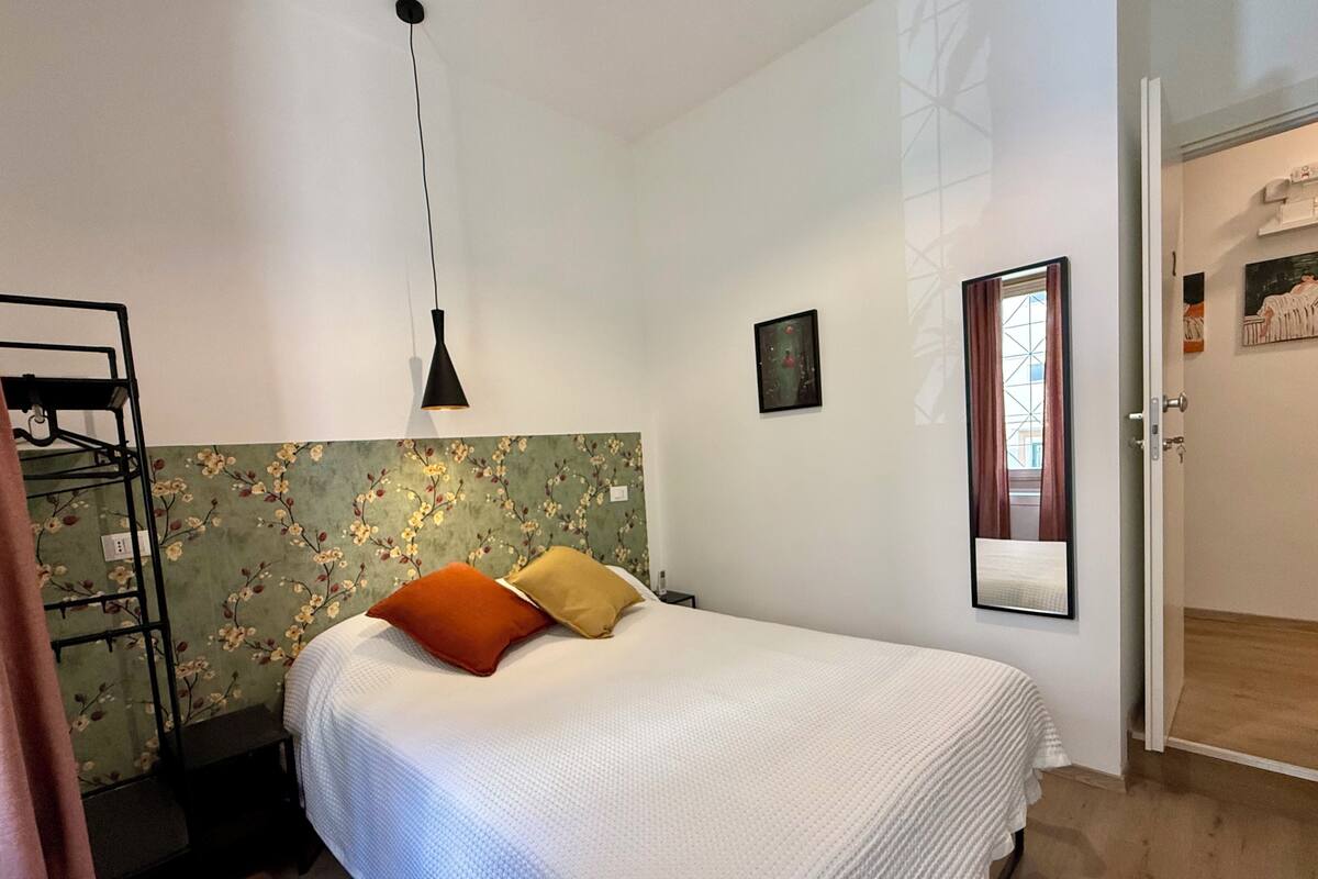 A bedroom with a bed, a hanging light, a floral wallpaper headboard, a painting, and a door leading to another room.