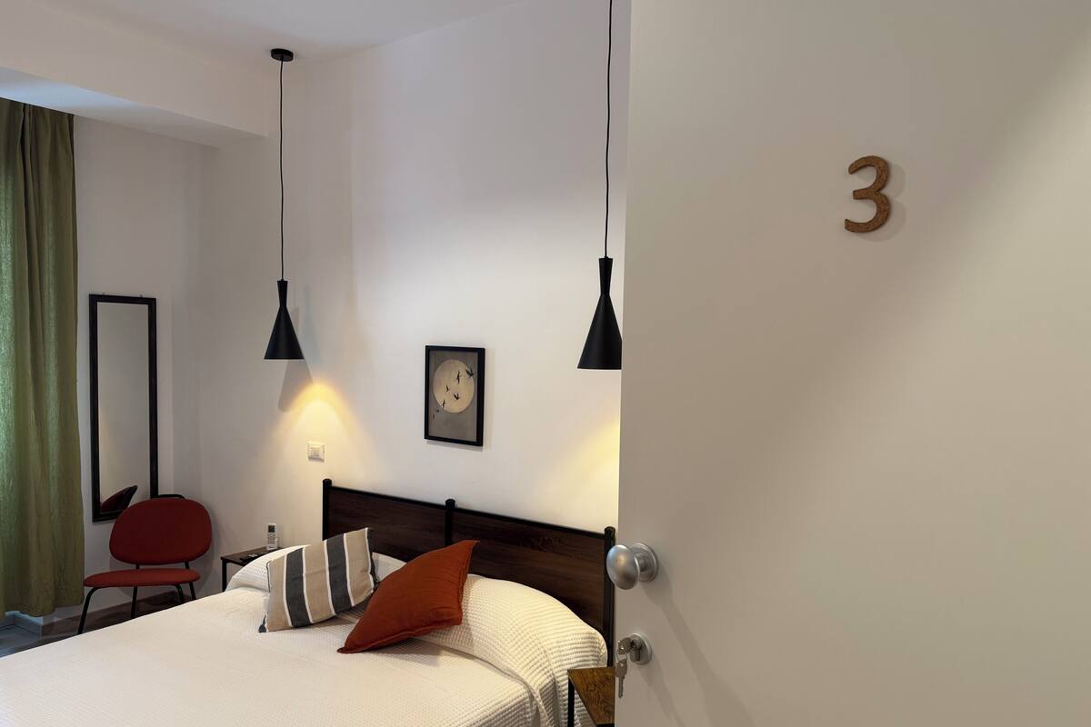 A hotel room with a bed, a chair, a bedside table, a wall clock, and a door marked with the number 3.