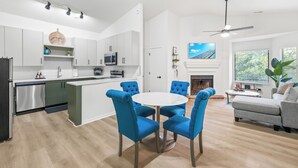 Una cocina moderna con armarios blancos, un microondas integrado y un refrigerador de acero inoxidable. Hay un comedor con una mesa redonda y sillas tapizadas en azul. Un sofá gris y una chimenea se ven en la sala de estar.