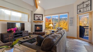 A cozy living room with a fireplace, a large window with a view of autumn trees, and a television.