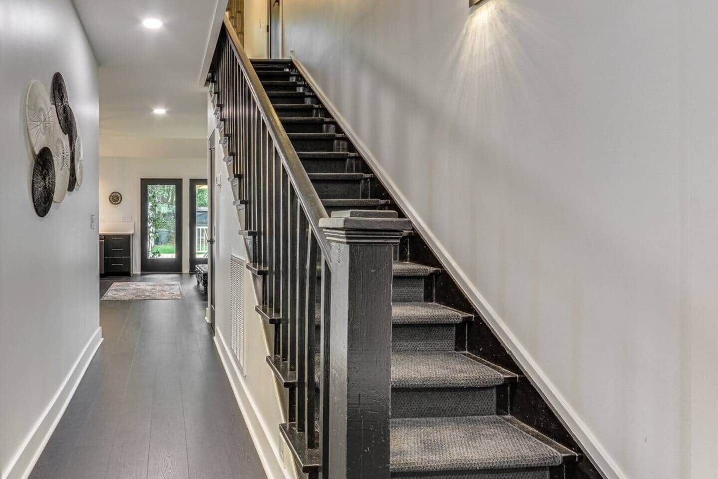 Un escalier moderne, doté d’une finition sombre et d’une main courante, mène à une pièce au plancher foncé et à la porte ornée d’un panneau de verre.