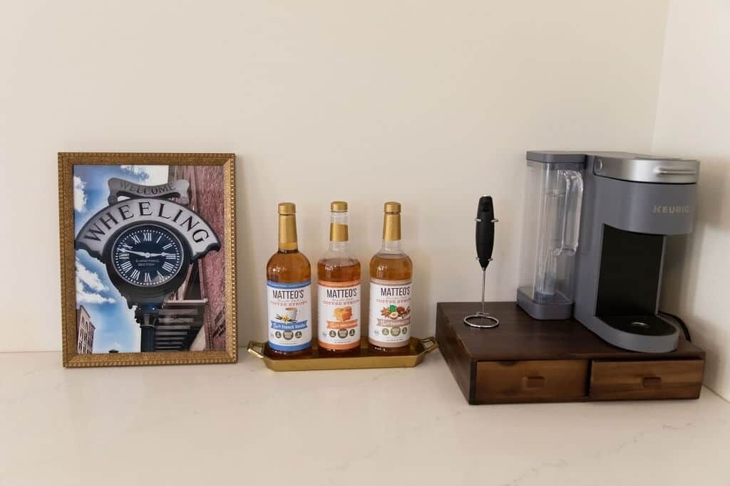 Eine gerahmte Uhr mit der Aufschrift 'WHEELING' und 'WELCOME', drei Flaschen Matteo's, ein schwarzer Stift und eine Keurig-Kaffeemaschine auf einem Holztablett.