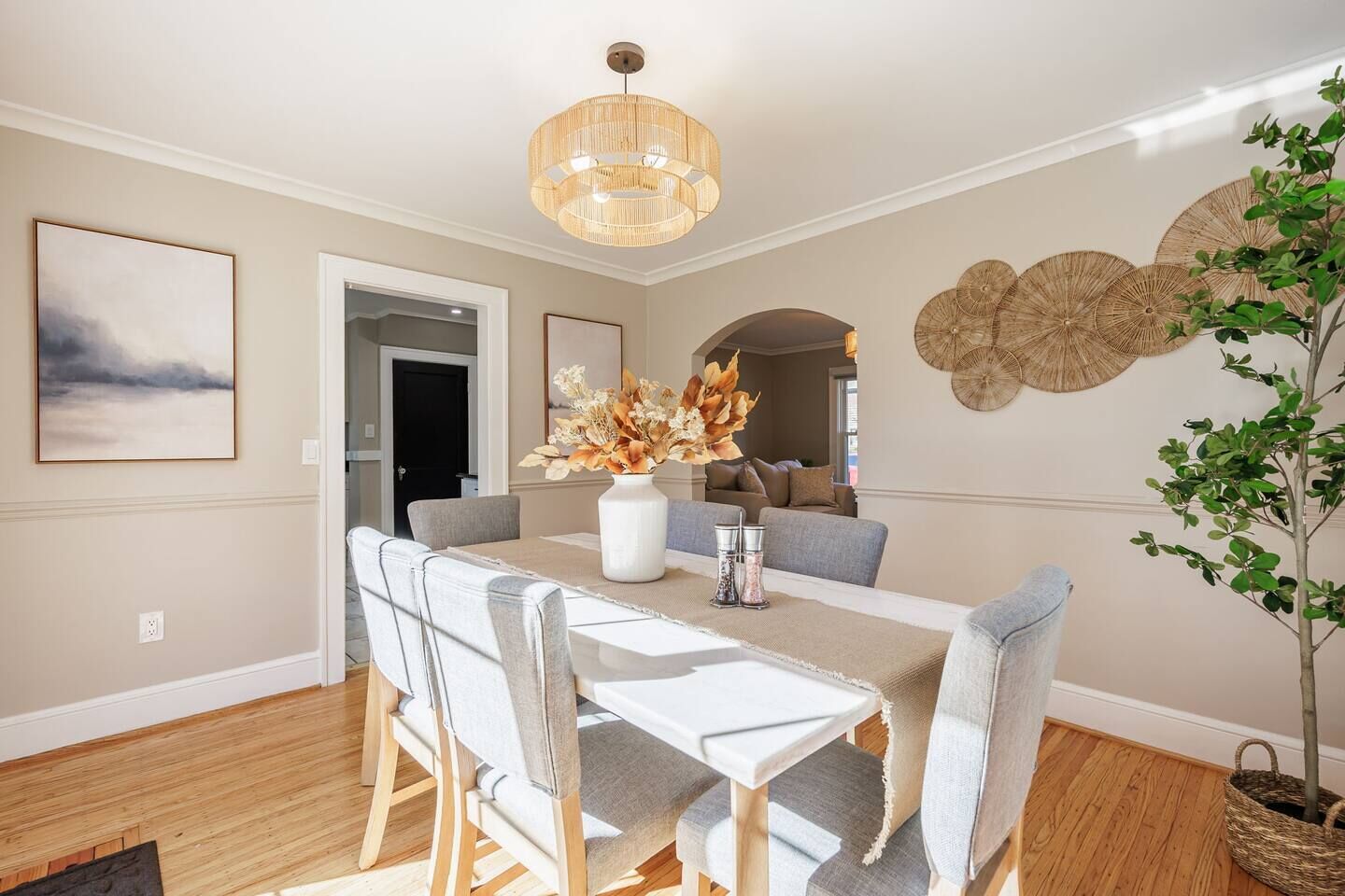 A dining area with a wooden table, grey upholstered chairs, a white vase with flowers, and a hanging light fixture.