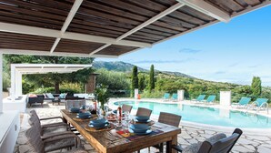 Mesa de comedor de madera dispuesta para seis personas, con sillas de mimbre, una piscina y sillones de descanso al fondo.