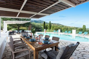 A wooden dining table set for six with wicker chairs, a pool, and lounge chairs in the background. - Thea Estate (Kassopei)