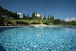 Une piscine à l’eau claire, entourée d’un jardin aménagé avec des arbres et des arbustes, et une colline sur laquelle se trouve une maison au loin.