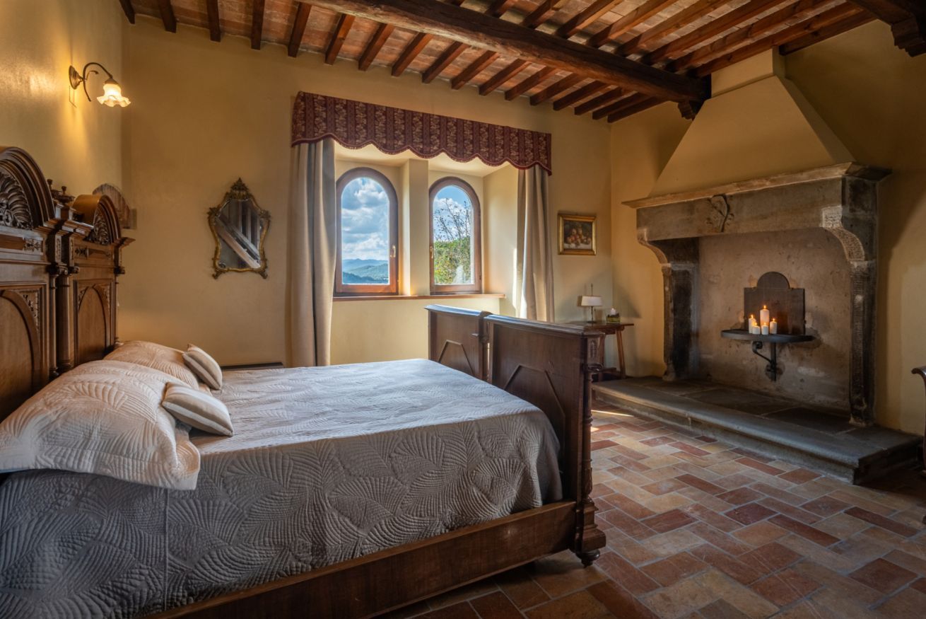 A bedroom with a bed, a fireplace, a window with curtains, and a stone floor.
