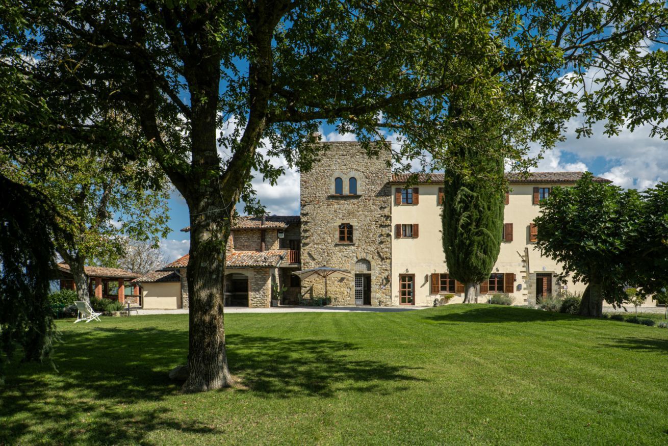 Un bâtiment en pierre, avec un grand arbre au premier plan, une pelouse verdoyante et un ciel dégagé.