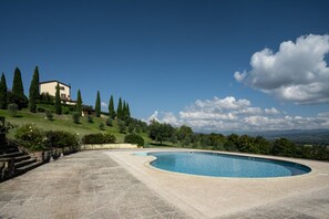 Una piscina de borde curvo, rodeada por un área empaveda, con una casa y árboles al fondo.