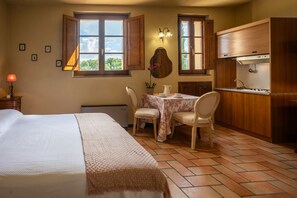 A bedroom with a bed, a desk with two chairs, a window with open shutters, and a kitchenette with wooden cabinets.