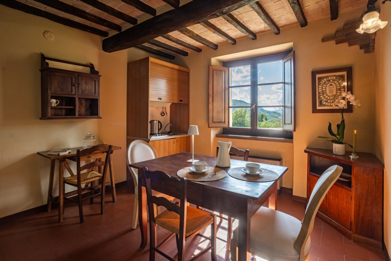 A dining area with a wooden table and chairs, a window with a view, and a kitchen area with wooden cabinets and a hanging pot rack.