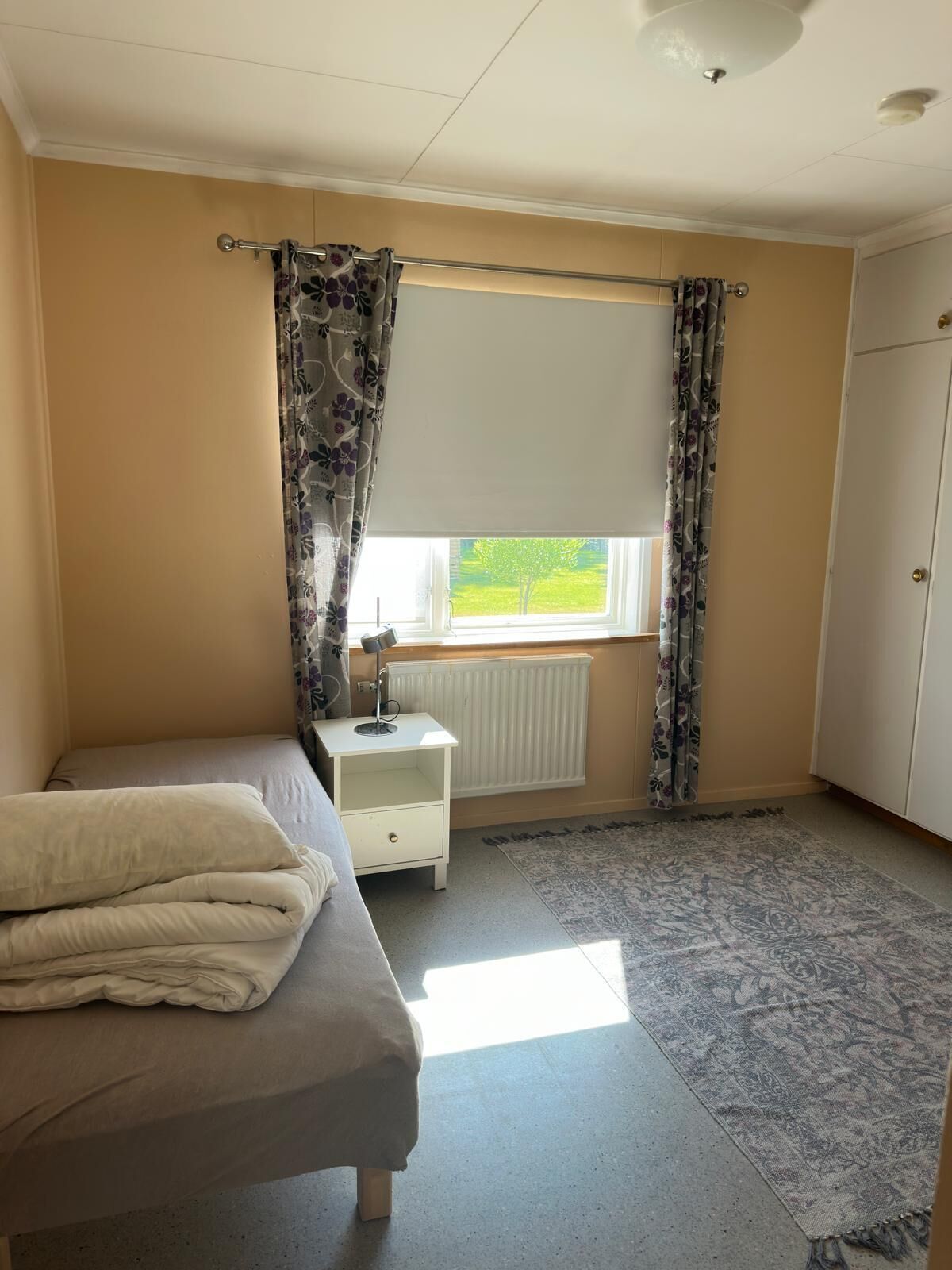 A bedroom with a bed, a nightstand, a radiator, a window with curtains, and a wardrobe.