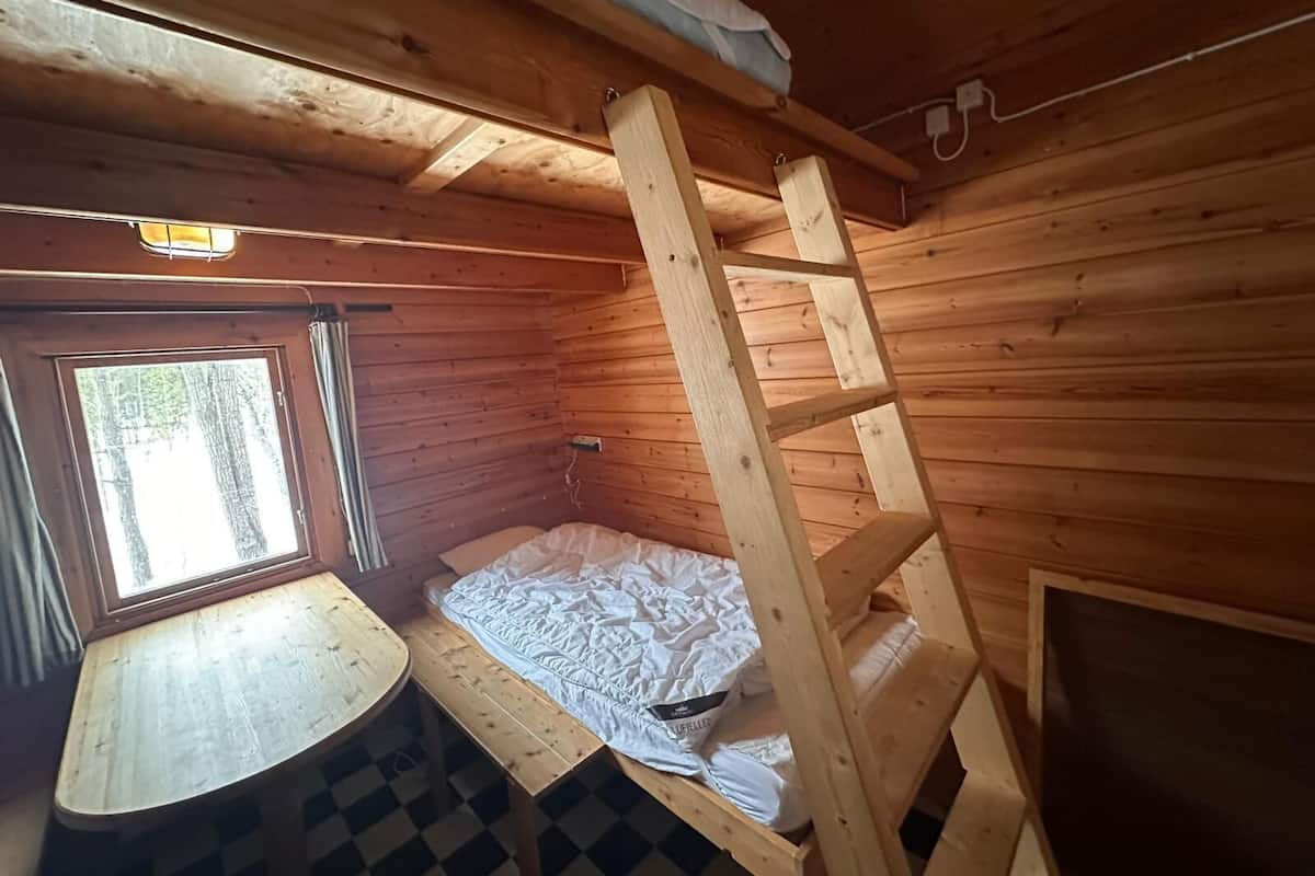 Ein kleines Zimmer in einer Holzhütte mit einem Etagenbett, einem Fenster, einem kleinen Tisch und einem karierten Boden.