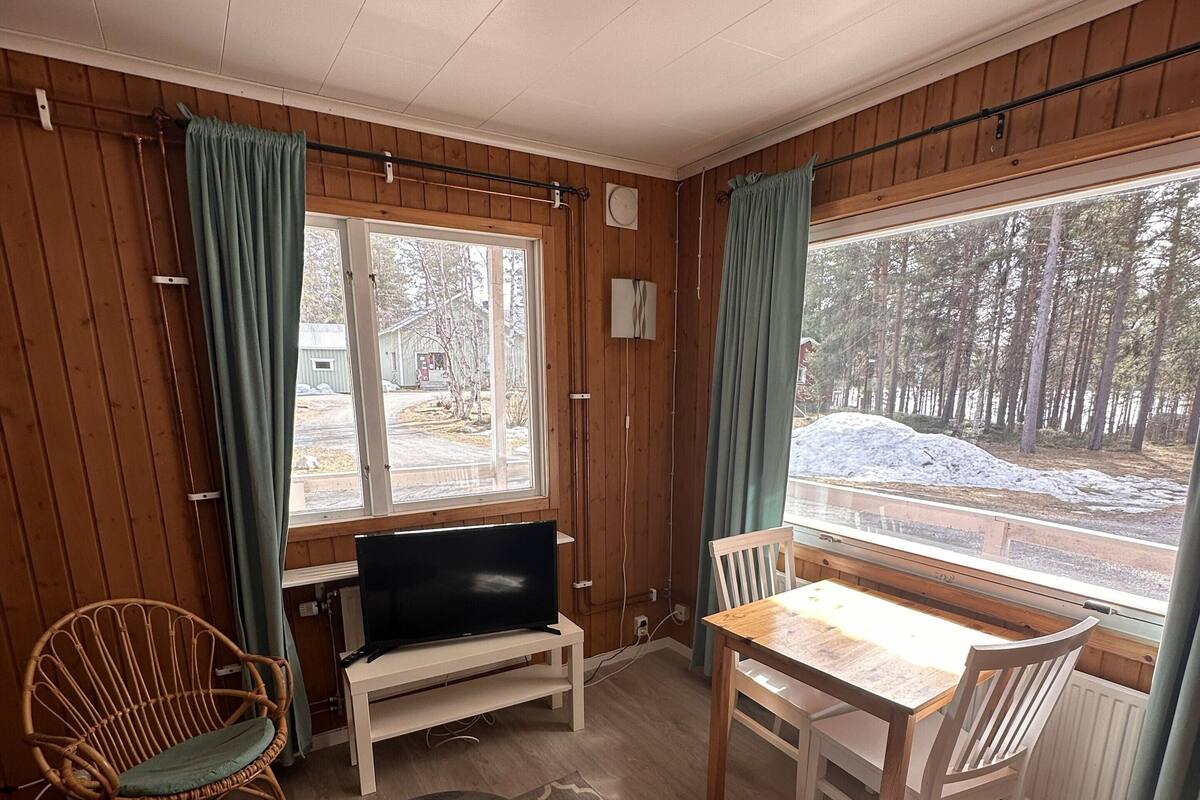 Ein Zimmer mit Holzwänden, einem Fernseher auf einem Standfuß, einem Esstisch mit Stühlen, einem gemusterten Teppich und großen Fenstern mit Vorhängen.