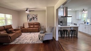 A spacious living room with a brown leather sofa, a grey armchair, and a patterned rug. A kitchen with white cabinets and stainless steel appliances is visible in the background.