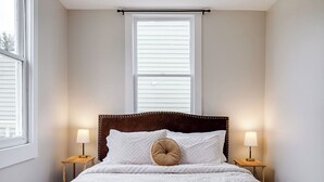 Une chambre avec une grande fenêtre, un lit avec une literie blanche, deux tables de chevet avec des lampes et une tête de lit en bois.