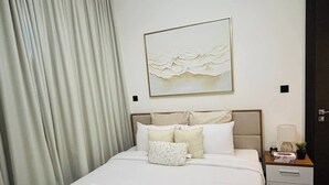 A neatly made bed with white linens and towels, a headboard with a painting, a bedside table with a lamp, and a large window with curtains.